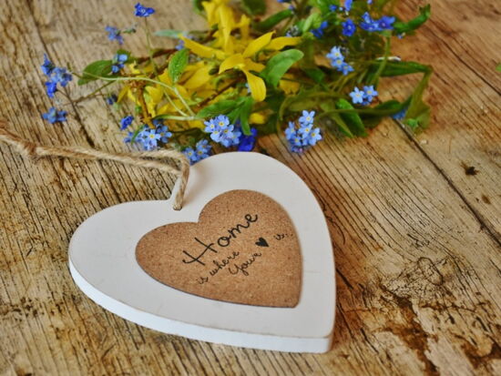 Zuhause ist, wo das Herz ist. Ein Holz- / Korkherz mit der Aufschrift "Home is where your heart is" übersetzt "Zuhause ist, wo das Herz ist". Es liegt auf einem Holztisch mit Wildblumen. Symbolbild für Wohnen und Heim.Holztisch