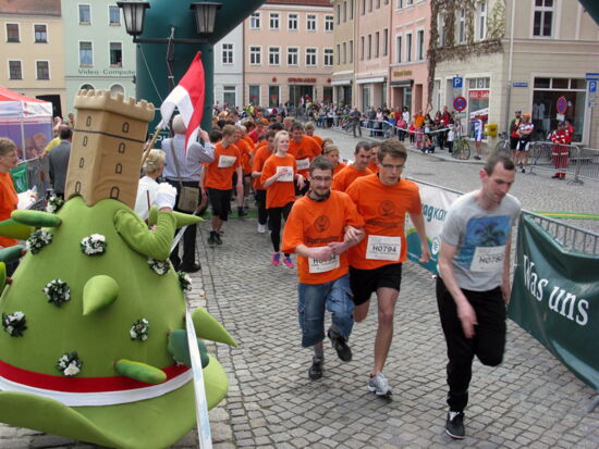 Lausitzer Blütenlauf - Inklusionslauf Laufrennstrecke des Kamenzer Blütenlaufes. Zahlreiche Teilnehmer, z.T. mit körperlicher Beeinträchtigung, laufen die Strecke entlang. Einige von ihnen laufen mit einem Partner. Sie tragen Jägermeister-T-Shors und Startnummern. Links Kami-Maskottchen.